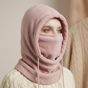 May include: A light pink balaclava-style hood with a face covering. The hood is made of a soft, fleece-like material and features a drawstring. The person in the image is wearing a cream-colored sweater.