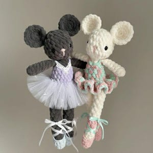 May include: Two crocheted mouse dolls dressed as ballerinas. One mouse is gray with a purple bodice and white tutu, the other is white with a pink and green bodice and tutu. Both wear ballet slippers and have large, round ears.