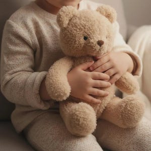Puede incluir: Un oso de peluche beige con una nariz marrón y ojos negros. El oso se sostiene en el regazo de alguien, su suave pelaje es visible. Los brazos y las piernas del oso son redondeados y tiene un diseño clásico de oso de peluche. Un juguete reconfortante.