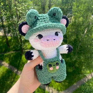 Puede incluir: Un peluche de vaca de ganchillo hecho a mano que lleva un sombrero y un peto de rana verdes. El peluche es principalmente blanco con pezuñas y detalles de orejas negros, un hocico rosa y ojos negros.