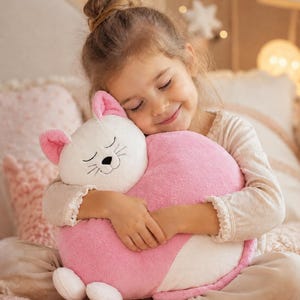 May include: A plush cat-shaped cushion featuring a white face with embroidered closed eyes, a black nose, and whiskers, paired with a soft pink body. The stuffed toy has small pink-lined ears and two white paws at the bottom, crafted from a textured fabric.