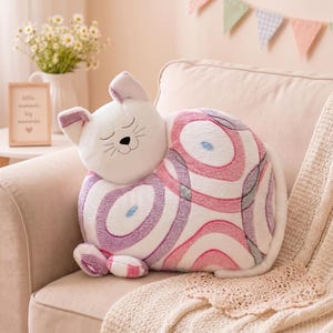 Puede incluir: Un cojín de peluche con forma de gato que presenta una cabeza blanca con ojos cerrados y bigotes bordados. El cuerpo es blanco con grandes patrones circulares y ovalados en tonos rosa, morado y gris. Tiene pequeñas patas y una cola.