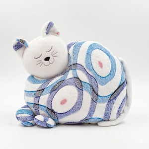 May include: A plush cat toy featuring a white head with embroidered closed eyes, a black nose, and whiskers. The body is covered in a soft fabric with a repeating pattern of blue, purple, and grey concentric circles on a white background.
