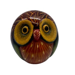 May include: A round, hand-painted wooden owl figurine featuring large, prominent yellow eyes with black pupils, a red beak, and a dark brown head with etched cross-hatch patterns. The chest is painted in shades of orange and red with white accents.