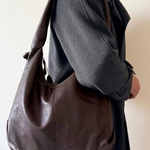 May include: A dark brown leather shoulder bag with a curved design. The bag has a long strap and a soft, slouchy appearance. The bag is worn over the shoulder, and the strap is a matching dark brown colour.