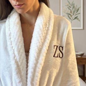 Może przedstawiać: Pluszowy, biały szlafrok z kołnierzem szalowym i pasującym paskiem. Szlafrok ma wyhaftowane inicjały "ZS" ciemnobrązową nicią na piersi. Miękka, puszysta faktura sugeruje komfort i relaks.