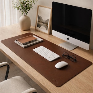 Puede incluir: Un tapete de escritorio de cuero marrón con un teclado, un ratón y gafas blancos. Un cuaderno y una foto de paisaje enmarcada también están en el escritorio. Un monitor de ordenador y un jarrón con vegetación completan el espacio de trabajo.