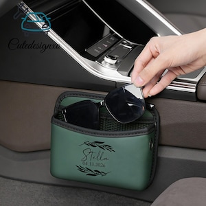 May include: A green car sunglasses holder with a black trim, holding a pair of sunglasses. The holder has the name "Stella" and the date "04.13.2026" printed on it. The holder is placed in a car.