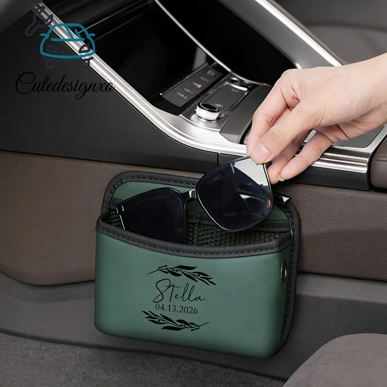 May include: A green car sunglasses holder with a black trim, holding a pair of sunglasses. The holder has the name "Stella" and the date "04.13.2026" printed on it. The holder is placed in a car.