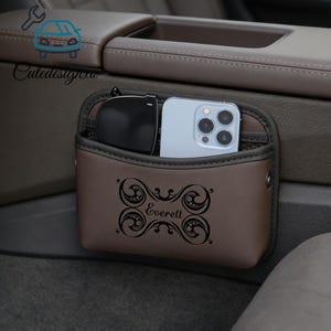 May include: A brown car organizer with a decorative design and the name "Everett" printed on the front. The organizer holds a pair of sunglasses and a smartphone. The organizer is designed to attach to a car's center console.