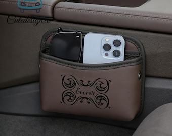 Può includere: Un organizer per auto marrone con un design decorativo e il nome "Everett" stampato sul davanti. L'organizer contiene un paio di occhiali da sole e uno smartphone. È progettato per essere fissato alla console centrale dell'auto.