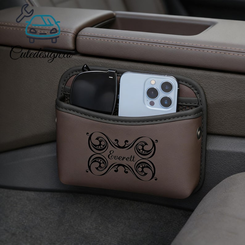 May include: A brown car organizer with a decorative design and the name "Everett" printed on the front. The organizer holds a pair of sunglasses and a smartphone. The organizer is designed to attach to a car's center console.
