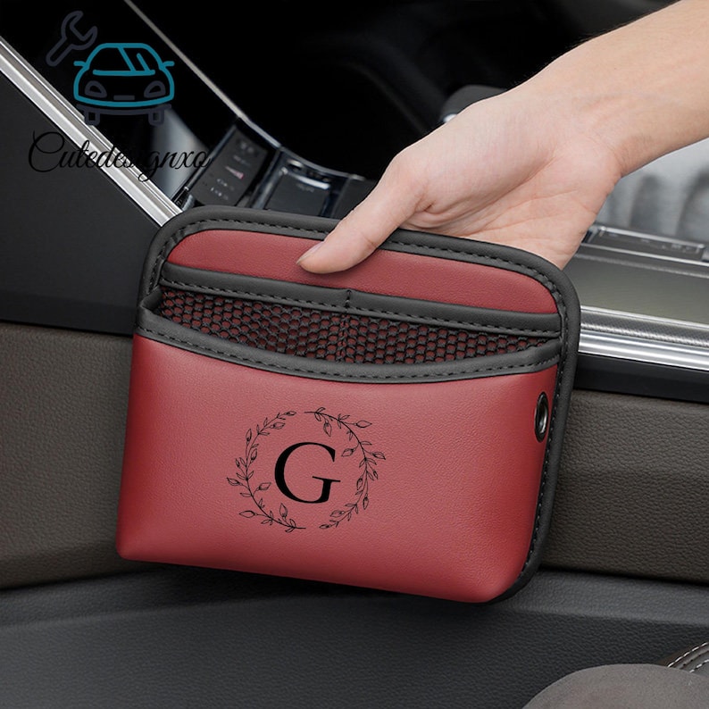 May include: A burgundy car organizer with a black mesh pocket and trim. The organizer features a black floral wreath design around the letter "G". The product is designed to be placed in a car.