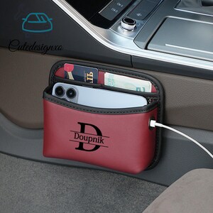 May include: A burgundy car organizer with a black trim, holding a smartphone, cards, and cash. The organizer has the name "Doupnik" and a "D" initial printed on it. A white charging cable is plugged into the organizer.