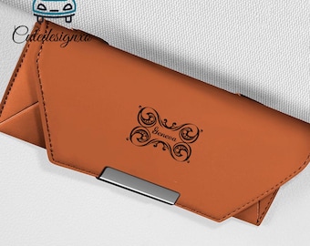 Può includere: Un portafoglio in pelle marrone con chiusura argentata. Il portafoglio presenta un design decorativo nero con il nome "Geneva" stampato sopra. Il portafoglio è chiuso e sembra nuovo.