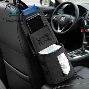 May include: Black car seat organiser with multiple compartments for storage. Features a phone pocket, tissue dispenser, and a blue card holder. The organiser is hanging from the headrest of a car seat. The word "Keenan" is printed on the organiser.