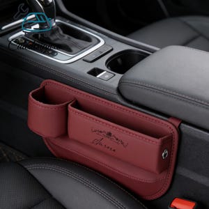 May include: A burgundy car seat organiser with multiple compartments, including a cup holder and two rectangular pockets. The word "Aurora" is printed on the front. The organiser is designed to fit between the car seats.