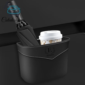 May include: Black car organizer with white stitching, holding a black umbrella and a coffee cup. The organizer has the name "Everett" and the letter "E" printed on it. The organizer is designed to fit in a car.