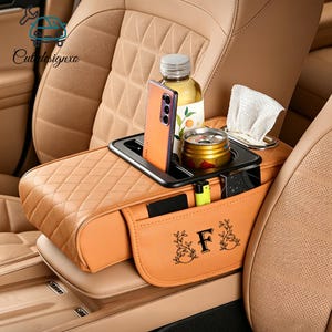 May include: Tan car console organizer with a black tray holding a phone, bottle, and can. The organizer has a quilted design and a pocket with the letter "F" and floral accents. A lighter and other items are in the pockets.