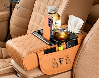 Può includere: Organizer per console auto color cuoio con un vassoio nero che contiene un telefono, una bottiglia e una lattina. L'organizer ha un design trapuntato e una tasca con la lettera "F" e decorazioni floreali.