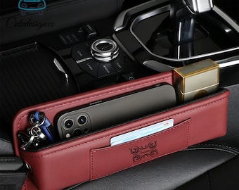 Può includere: Un organizer per sedili auto bordeaux con più scomparti. Contiene uno smartphone, chiavi, un accendino e una carta. L'organizer è progettato per adattarsi tra il sedile e la console centrale.