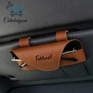 May include: A brown leather car visor sunglasses holder with the name "Forrest" printed on the front. The holder is attached to the car's visor with two straps. A pair of glasses is partially visible inside the holder.