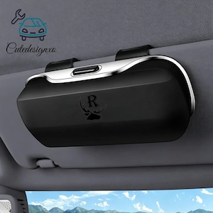 May include: A matte black rectangular car visor sunglasses holder with a silver metallic top trim and a central push-button release. The front features a debossed logo of a deer head silhouette with the letter R above it, mounted on a gray visor.