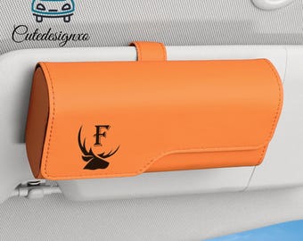 Può includere: Un organizer per aletta parasole per auto arancione con una testa di cervo nera e la lettera "F" stampata sul davanti. L'organizer è rettangolare e ha una cinghia per il fissaggio all'aletta parasole dell'auto. Il logo "Cutedesignxo" è nell'angolo in alto a sinistra.