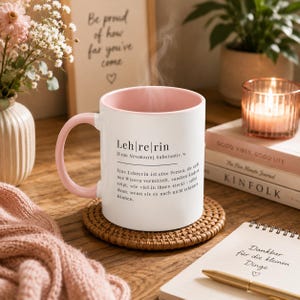 May include: A white ceramic mug with a soft pink interior and handle, featuring black printed German text that defines the word Lehrerin. The mug sits on a round woven coaster, with steam rising from the top, indicating a hot beverage inside.