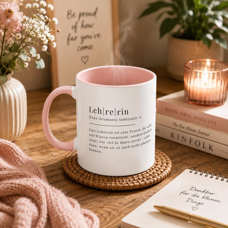 May include: A white ceramic mug with a soft pink interior and handle, featuring black printed German text that defines the word Lehrerin. The mug sits on a round woven coaster, with steam rising from the top, indicating a hot beverage inside.