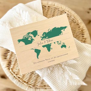 May include: A wooden world map with green continents and the names "Julia & Tobias" printed in black. Below, the text reads "Die sch&ouml;nste Reise ist die gemeinsame 14.06.2026". The map is set on a white blanket and a wicker basket.