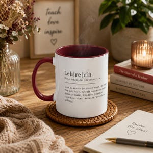 May include: A white ceramic mug with a deep red interior and handle, featuring black printed German text that defines the word Lehrerin. The mug sits on a round woven coaster, with steam rising from the top, displaying a dictionary-style entry.