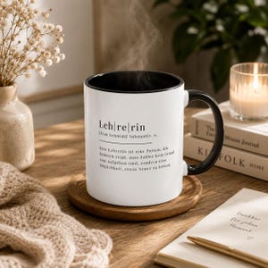 May include: A white ceramic mug with a black interior and handle, featuring black printed text that defines the word Lehrerin, including the name Frau Schmidt, with steam rising from the top of the mug while it sits on a round wooden coaster.