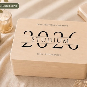 Könnte beinhalten: Eine rechteckige, helle Holzbox mit Klappdeckel. Auf der Oberseite befindet sich der schwarze Schriftzug "DEIN ABENTEUER BEGINNT.", die Jahreszahl "2026" mit dem mittig platzierten Wort "STUDIUM" sowie der Name "LENA" und "PSYCHOLOGIE".