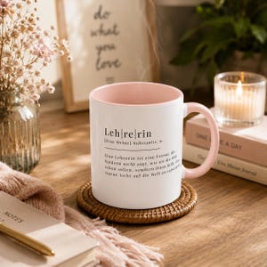 May include: A white ceramic mug with a soft pink interior and handle, featuring black printed German text that defines the word Lehrerin. The mug sits on a round woven rattan coaster, with steam rising from the top, displaying a dictionary-style entry.