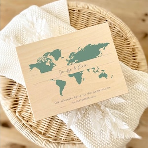 May include: A wooden world map with teal continents and the names "Jeniffer & Kevin" inscribed. Below, the text reads "Die sch&ouml;nste Reise ist die gemeinsame 20. SEPTEMBER 2026". The map is set on a cream-colored blanket inside a wicker basket.