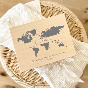May include: A wooden world map with gray continents and the names "Laura & Marcel" in cursive. Below, the text reads "Die sch&ouml;nste Reise ist die gemeinsame 31.MAI 2026". The map is set on a white blanket and a wicker basket.