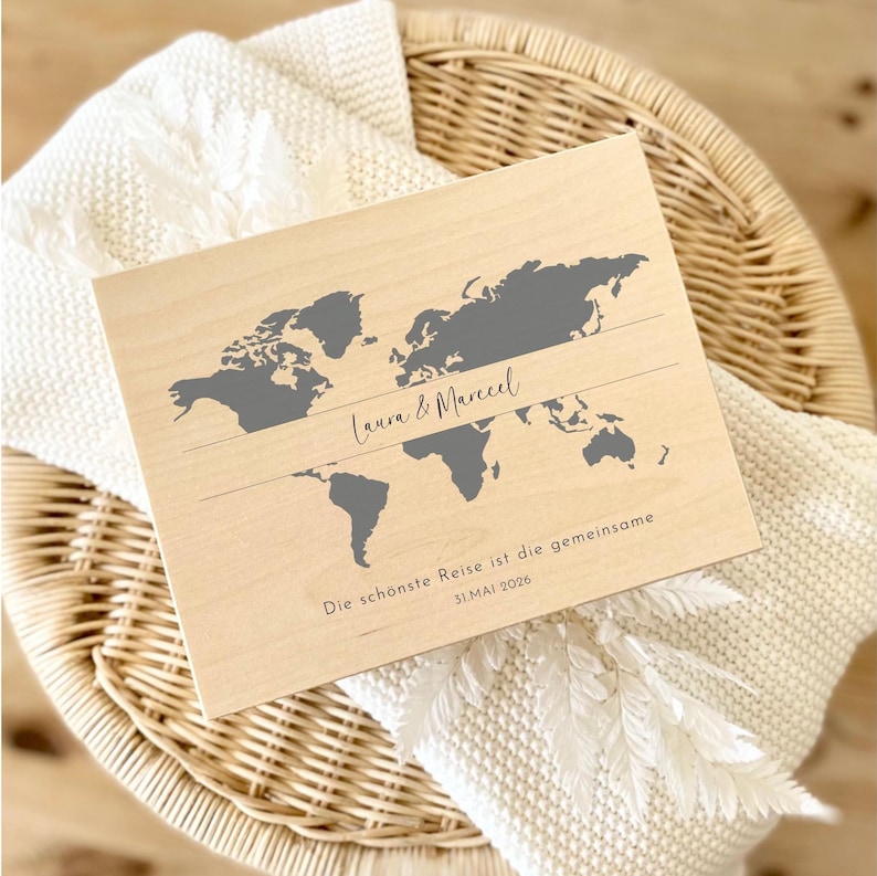 May include: A wooden world map with gray continents and the names "Laura & Marcel" in cursive. Below, the text reads "Die sch&ouml;nste Reise ist die gemeinsame 31.MAI 2026". The map is set on a white blanket and a wicker basket.
