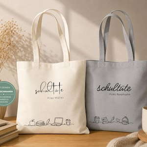 K&ouml;nnte beinhalten: Zwei Tragetaschen aus Canvas, eine cremefarbene und eine graue, mit schwarzer Schrift "Schult&uuml;te" und einem Namen darunter. Die cremefarbene Tasche hat eine Strichzeichnung von Schulmaterial. Die graue Tasche hat ein &auml;hnliches Design. Beide haben lange Henkel.