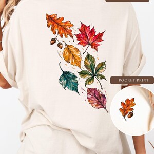 May include: A cream-colored t-shirt featuring a large back print of watercolor-style autumn leaves in shades of orange, red, yellow, green, and purple, accompanied by two small acorns. A circular inset shows a smaller matching pocket print.
