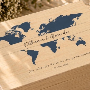 May include: Wooden box with a world map design in navy blue. The box is engraved with the names "Katharina & Alexander" and the date "31.MAI 2026". The text "Die sch&ouml;nste Reise ist die gemeinsame" is also engraved on the box. The box is made of light-colored wood.