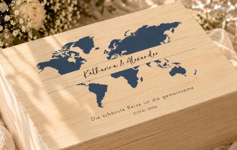 May include: Wooden box with a world map design in navy blue. The box is engraved with the names "Katharina & Alexander" and the date "31.MAI 2026". The text "Die sch&ouml;nste Reise ist die gemeinsame" is also engraved on the box. The box is made of light-colored wood.