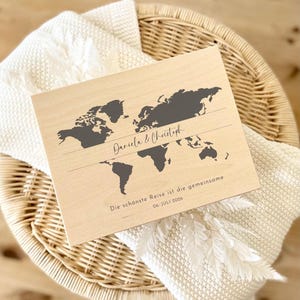 May include: A wooden world map with a dark gray outline of the continents. The names "Daniela & Christoph" are written above a horizontal line. Below the line, the text reads "Die sch&ouml;nste Reise ist die gemeinsame 06. JULI 2026".