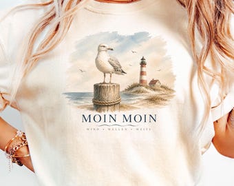 Può includere: Una t-shirt color crema con una grafica in stile acquerello che raffigura un gabbiano appollaiato su un palo di legno vicino a un faro e all'oceano. Il testo MOIN MOIN è stampato sotto la grafica, seguito dalle parole WIND • WELLEN • WEITE.