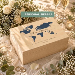 Könnte beinhalten: Eine hellfarbene Holzkiste mit blauem Weltkarten-Design und den Namen "Katharina & Alexander". Der Text "Die schönste Reise ist die gemeinsame" ist ebenfalls sichtbar. Die Kiste ist von Blumen und Kerzen umgeben.
