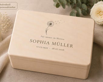 Può includere: Una scatola rettangolare in legno chiaro con un disegno a linee nere di un dente di leone con i semi che volano via. Il testo "Für immer im Herzen", "SOPHIA MÜLLER" e le date "12.05.1945" e "28.07.2026" sono stampati in inchiostro nero.