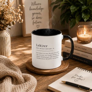 May include: A white ceramic mug with a black interior and handle, featuring the German word Lehrer defined with text in black ink. Steam rises from the mug, which sits on a round wooden coaster. The mug displays a dictionary-style definition.