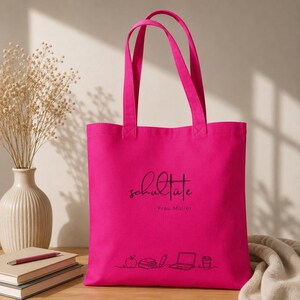 K&ouml;nnte beinhalten: Eine leuchtend pinkfarbene Tragetasche mit schwarzem Text, der "Schult&uuml;te Frau M&uuml;ller" lautet. Die Tasche hat zwei Henkel und eine Strichzeichnung eines Apfels, B&uuml;cher, eines Bleistifts, eines Laptops und einer Kaffeetasse.