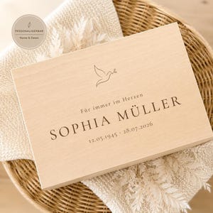 Könnte beinhalten: Eine rechteckige, helle Holzschatulle mit einer Lasergravur einer Taube, die einen Olivenzweig trägt. Der Text lautet "Für immer im Herzen", "SOPHIA MÜLLER" sowie die Daten "12.05.1945 - 28.07.2026" in einer dunkelbraunen Schrift.