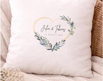 Può includere: Cuscino quadrato bianco con un design a forma di cuore dorato, ornato da foglie verdi e blu. I nomi "Julia & Tobias" e la data "16 agosto 2026" sono scritti all'interno del cuore. Il testo "PERSONALISIERBAR" è in alto.